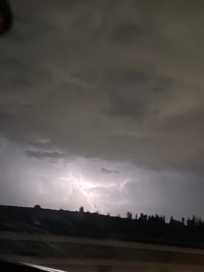 The image displays a nocturnal storm scene, attributed to the Megido Regional Council, Israel. The sky is predominantly filled with dark, dense storm clouds. A prominent, bright white-blue lightning bolt, appearing as a multi-branched electrical discharge, is centrally positioned in the lower-mid portion of the frame, illuminating the surrounding cloud mass. Below the illuminated clouds, a dark, silhouetted landform, consistent with a low ridge or hillside, extends horizontally across the lower mid-ground. Several indistinct dark tree silhouettes are discernible along the crest of this landform. The immediate foreground is dark and unidentifiable, showing only faint, horizontal textural variations. No individuals, human activity, or discernible man-made structures are present within the frame.