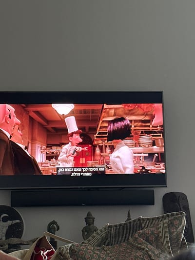 The image displays an indoor setting with a flat-screen television centered in the mid-ground. The television screen presents an animated scene from the film "Ratatouille," featuring three primary characters within a professional kitchen environment. On the left, two male figures in dark suits are partially visible. In the center, Linguini, adorned in a tall chef's hat and white chef's coat, holds a small dark object in his left hand and points right with his right hand, appearing to speak. To his right, Colette, with short dark hair and a white chef's coat, faces Linguini. The kitchen setting includes stainless steel surfaces, various pots, pans, and shelves, under a single overhead light fixture. Hebrew subtitles are displayed at the bottom of the screen, reading: "הוא הסיבה לכך שאנו נמצאו כאן, מאחורי הדלת." (He is the reason we are here, behind the door.)
Directly beneath the television is a horizontal black soundbar. Below the soundbar, on a surface, several objects are arranged. From left to right: a dark circular plaque with a white outline of the Vitruvian Man, followed by three distinct dark figurines, two of which resemble stylized Asian religious figures. A large, patterned fabric object, featuring floral and striped designs with visible red accents, occupies the right foreground, partially obscuring the lower portions of the figurines. The upper portion of the image shows a plain, light gray wall above the television. The specific geographical location in Merom HaGalil Regional Council, Israel, cannot be determined from the visual content of this image.