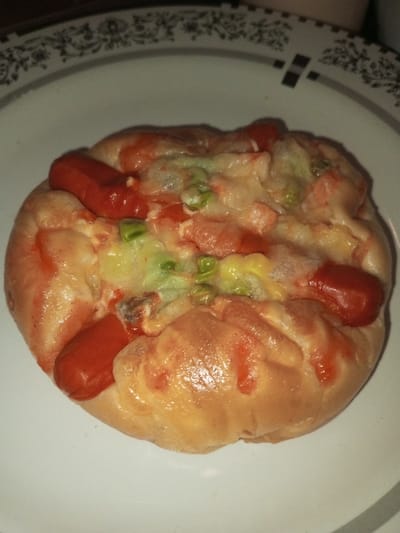 A round, baked food item, approximately 15 cm in diameter, is centered on a white plate with a decorative rim featuring dark gray/black floral and geometric patterns. The food item consists of a light golden-brown dough base, topped with three reddish-orange mini sausages embedded around the perimeter. The center is covered with melted white and yellow cheese, green peas, and diced red ingredients, possibly tomatoes or bell peppers. Red sauce, consistent with ketchup or chili sauce, is drizzled over the toppings and dough. The scene depicts a prepared meal or snack indoors in Khajuripada, India.