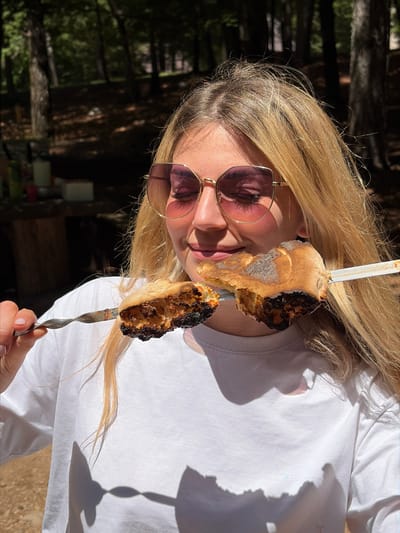 A female subject is depicted outdoors in a densely wooded area in Mali Bostur, Montenegro, under direct sunlight. She is positioned centrally, from the mid-torso upwards, with her head slightly tilted. The subject has long, light blonde hair with visible individual strands and wears large, hexagonal-shaped sunglasses with pink-tinted lenses and thin gold frames, obscuring her eyes. Her mouth is slightly curved in a subtle smile. She is wearing a plain white, long-sleeved crew-neck t-shirt.

She holds two distinct metal skewers, each impaling a piece of cooked food, directly in front of her face. The food items are irregular in shape, appearing bread-like or dough-based, with significant areas of charring and dark brown to black coloration on the undersides and edges, and lighter golden-brown, possibly sugary, surfaces on top. The skewer held in her left hand (viewer's right) has a twisted metal handle design. The skewer held in her right hand (viewer's left) appears to have a flat, blade-like shaft.

The background consists of numerous tall, dark tree trunks and green foliage, indicating a forest or wooded environment, with strong dappled sunlight and deep shadows across the scene. On the blurry left side of the background, indistinct objects resembling a red cup, a white box, and dark green containers are visible on a surface, suggesting an outdoor picnic or gathering. Prominent shadows from the subject's head, hair, and the skewers are cast onto her white shirt.