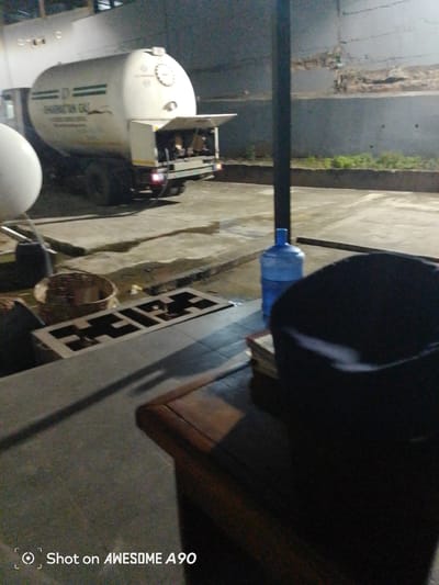 In the city of Ibadan, Nigeria, an outdoor scene at night depicts a white gas tanker truck with "BHARATTAN GAS" written on its side. The truck is parked on a concrete surface, next to a large white gas cylinder. In the foreground, a dark-colored woven basket sits on the concrete, near a metal grate. Further to the right, a large blue water dispenser jug is visible, and beyond that, a dark object, possibly a chair or bag, is partially seen. The background features a grey wall and some vegetation. The overall impression is a low-light, utilitarian setting. The bottom left corner of the image contains a watermark that reads "Shot on AWESOME A90".