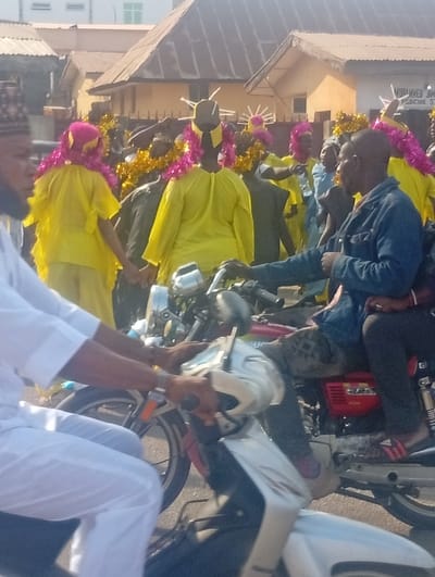 This image captures a daytime street scene in Ozuri, Nigeria, featuring a group of people, some on motorcycles, set against a backdrop of urban buildings.
In the left foreground, a man wearing a white traditional outfit and a dark cap is partially visible, operating a light-colored scooter with his hands on the handlebars. To his right, a second man in a blue jacket and dark pants is seated on a red motorcycle, identifiable by a "CD 50" emblem on its side, looking towards the left. A passenger, wearing dark clothing and pink-striped sandals, sits behind him on the same motorcycle.
In the midground, a procession or gathering of approximately ten individuals is observed. Most are dressed in bright yellow long-sleeved garments. Several members wear elaborate headwear made of pink tinsel and have golden tinsel garlands around their necks or shoulders. Some individuals display distinct dark, spiked headpieces resembling crowns or rays. Two people in yellow are holding hands, while another in the group appears to be pointing to the right.
The background consists of several single and multi-story buildings with corrugated metal roofs. A prominent sign on a building to the right is partially legible as "MUHAMMED JAM MEDICINE STORE." Further back, a multi-story building with light-colored walls and a green-framed window is visible. The lighting indicates clear, sunny conditions.