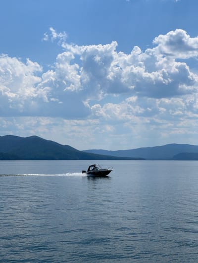 A motorboat is cutting through calm, blue water, leaving a foamy wake behind it. The boat is dark and sleek, with a small windshield and a canopy overhead. It appears to be moving at a good speed, as indicated by the spray it's kicking up. In the background, hazy blue mountains rise up from the water's edge, their slopes covered in dark greenery. The sky above is a brilliant blue, dotted with large, fluffy white clouds that are illuminated by the sun. The overall scene suggests a bright and pleasant day, perfect for a boat trip on the water. The image is oriented vertically, with the sky and clouds on the left and the water and boat on the right.
There is no visible text in the image.