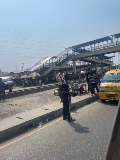 The scene depicts a busy street and market area in Festac Town, Nigeria, under a clear, bright sky. A paved asphalt road occupies the foreground, bordered by a concrete curb with visible drainage openings and scattered litter. A yellow solid line is painted on the road adjacent to the curb.

Beyond the curb, a wide, unpaved median strip, predominantly sandy and dusty, hosts numerous market stalls. These stalls are sheltered by various colored umbrellas and canopies, including green, red, and purple, and display goods on tables and directly on the ground. Litter, consisting of plastic bags and bottles, is present across this median.

Dominating the midground and background is a large concrete pedestrian overpass. This structure features multiple staircases and ramps, with metal railings and a translucent blue roof covering its walkways. Power lines are visible against the sky, crossing above the overpass.

Numerous individuals populate the scene. In the immediate foreground, a male figure wearing a black tracksuit with white side stripes stands on the road next to the curb, holding a dark rectangular object with both hands at waist level. His shadow is cast distinctly on the asphalt. On the far right, a yellow commercial minibus, identified by "F.C.B" and "LAGOS" markings, is partially visible, oriented towards the left.

On the median strip, multiple people are gathered around the market stalls. A male in a light-colored, patterned short-sleeved shirt and dark trousers stands near the center of the median, carrying a small dark shoulder bag. To his right, a group of individuals, including a male in a black and white striped shirt, are standing near the yellow minibus. A light-colored van is parked further left on the median. Additional people are visible on the pedestrian overpass, suggesting active use. Interactions appear to be typical of a bustling marketplace, involving transit, vending, and shopping.