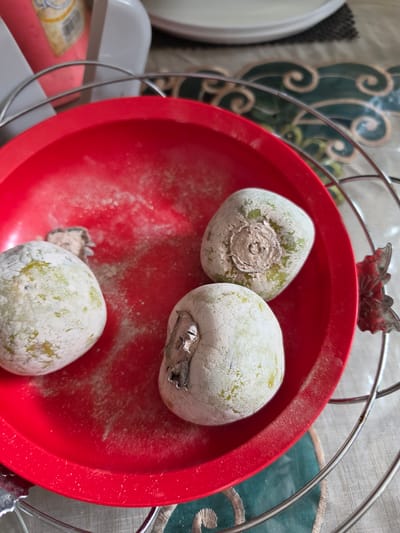 A close-up shot shows three round, pale green fruits, possibly persimmons, in a bright red bowl. The fruits are covered in a powdery white residue and have visible stem scars. The bowl sits on a patterned tablecloth with green and white swirling designs. A silver wire rack surrounds the bowl, with a decorative metallic accent resembling a leaf or flower on the right side. In the background, out of focus, are bottles and containers, one pink with a yellow label. The lighting suggests an indoor setting. There is no indication of the specific location within the city of Medan, Indonesia.