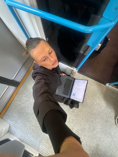 A woman is standing on a train with a laptop and tablet, looking down at her screen. She is wearing a black jacket and black pants. The train has blue railings and a gray floor. The location is not Tonbridge and Malling, United Kingdom.