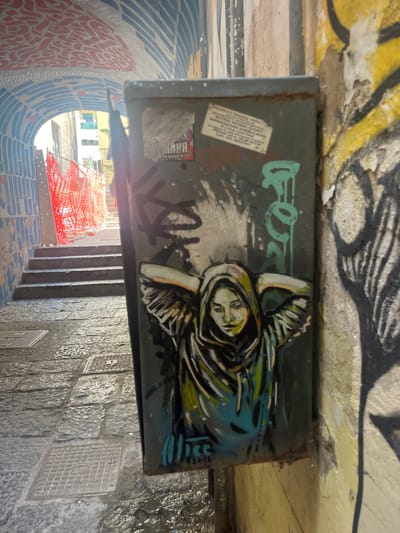 The image shows a narrow, sloped alleyway in Salerno, Italy. The walls are covered in graffiti and the ground is paved with cobblestones. In the foreground, a metal electrical box is plastered with various stickers and features a stencil of a person wearing a hooded sweatshirt. To the left, a set of stone stairs leads upwards, partially obscured by orange construction netting. The overall impression is one of urban decay and artistic expression.