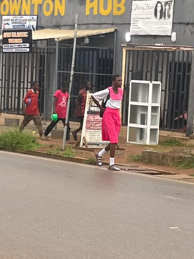The image shows a street scene in Sabonjibi, Nigeria, with a group of individuals walking on a sidewalk and crossing the street. In the foreground, a person is walking from right to left, wearing a white shirt with a pink collar and bib, and bright pink shorts. They have dark hair and are wearing white socks and sandals. Behind them, two other individuals are walking in the same direction. The person on the left is wearing a red shirt and brown pants, carrying a red object. The person in the middle is wearing a pink t-shirt and black pants, holding a green bowl. Another person in a red and orange striped shirt is visible further behind.

The background features a building with yellow signage that reads "DOWNTON HUB". Below the signage are various signs, including one for "CHRIS CABI LAUNDRY AND DRY CLEANING SERVICES" and another for "ICE CLASSIC WEARS". The building has a row of barred windows and a light-colored overhang. Adjacent to the building on the right is a large white object that appears to be a disassembled window frame or display unit. The ground is paved with asphalt, and there is a strip of green vegetation along the side of the road. The overall lighting suggests it is daytime.
