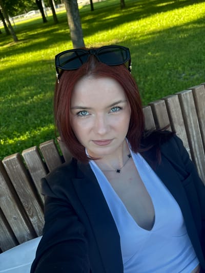 A woman with red hair is sitting on a park bench in Sinyavino, Russia. She is wearing black sunglasses on top of her head, a black blazer, and a white V-neck top. She is looking at the camera with her blue eyes. The background is a grassy park with trees and sunlight filtering through them. The bench is made of wooden slats.