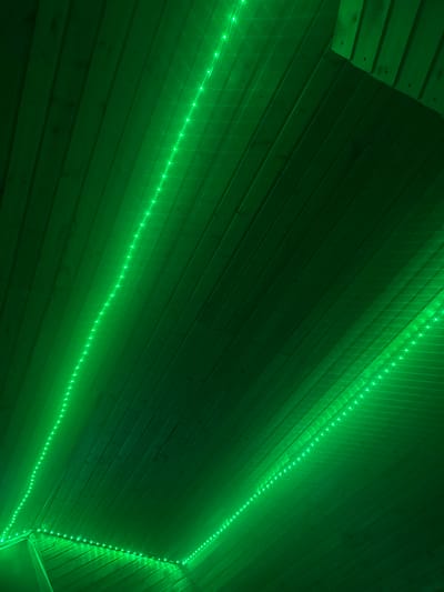 The image shows a wooden ceiling with green LED strip lights. The lights are installed along the edges of the ceiling panels, creating a vibrant green glow that illuminates the wooden surface. The wooden panels are oriented diagonally from the bottom left to the top right of the frame. There are no people or other discernible objects in the scene. The overall impression is of an indoor space decorated with modern lighting. The location in Семибратово, Russia, is not evident from the visual information provided in the image.