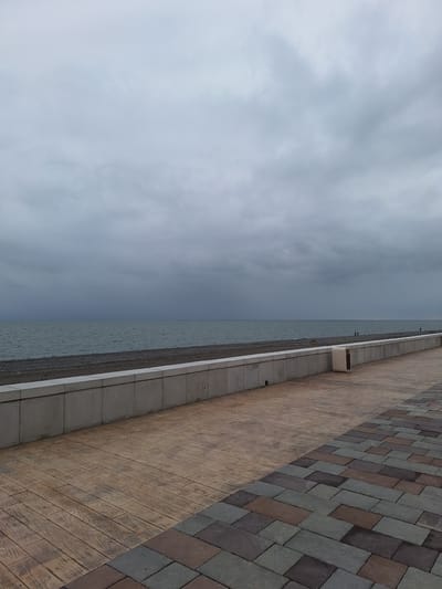 The image captures a section of the waterfront promenade in Kobuleti, Georgia, under an overcast sky. A wide, paved walkway dominates the foreground, transitioning from a wooden-patterned surface on the left to a multicolored brick pattern on the right. Adjacent to the walkway is a low, modern concrete barrier that separates it from the pebble beach and the dark, calm sea. In the distance, several indistinct figures are visible on the beach, and the horizon is marked by a blurred line where the sea meets the heavy, gray clouds. A single, light-colored trash receptacle is positioned along the concrete barrier. The overall atmosphere is somber and muted, indicative of a cloudy or pre-rainy day.