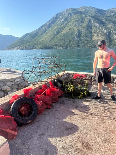 This image captures a sunlit coastal scene in Prčanj, Montenegro. An adult male stands on a concrete pier in the mid-ground, positioned toward the right side of the frame. He is shirtless, wearing black shorts, dark sneakers, a dark baseball cap, and sunglasses, with a pendant visible on a necklace. To his left, on the concrete surface, are various objects: two large, rusty, circular wire-frame structures leaning against each other, a rectangular frame covered in green netting, and a collection of red mesh bags, many containing spherical dark objects. A black rubber tire also rests among the red mesh bags. The pier edge to the left is a low stone wall topped with concrete. Beyond the pier, calm, blue-green water stretches across to the opposite shore, where a town's buildings are clustered at the base of steep, vegetated mountains. The sky is clear blue, with one small cloud visible on the left horizon. Strong sunlight casts distinct shadows across the pier surface.