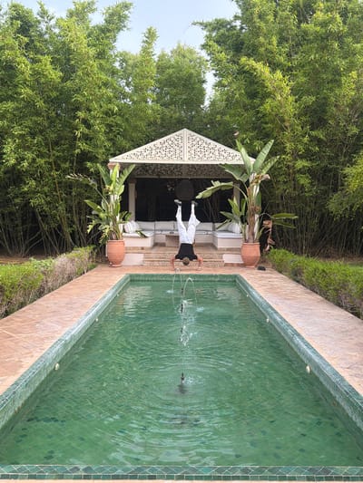 In Al Ouidane, Morocco, a person performs a handstand at the edge of a rectangular swimming pool. The pool has vibrant green tiled walls and a tiled bottom, with a fountain in the center creating ripples on the water's surface. The person is wearing white pants and a dark top, with their legs extended upwards. To the rear of the pool, a pathway of terracotta tiles leads to a white, ornate gazebo. This gazebo features a patterned roof and contains white seating. Flanking the gazebo are two large potted plants with broad leaves, and behind them, a dense wall of tall bamboo stalks forms a natural backdrop. The area is bathed in natural daylight.