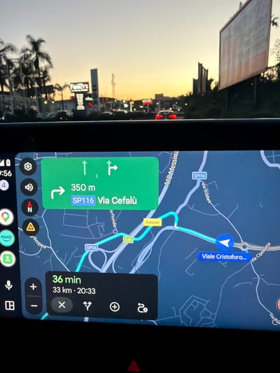 The image depicts a car's dashboard screen displaying a Google Maps navigation interface, indicating a route to Via Cefalù, a destination 350 meters away. The route is being navigated in Acireale, Italy. The screen also shows an estimated travel time of 36 minutes and a distance of 33 kilometers. The road network visible on the map includes SP116, A18 Messina, Viale Cristoforo and E45. The time on the dashboard is 9:56 PM and the estimated arrival time is 8:33 PM. Visible through the windshield is the street view of Acireale at sunset, where a Franzy’s shop sign can be seen.