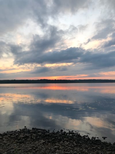 The image shows a serene lake at dusk in Svirstroy, Russia. The sky is filled with dramatic, gray clouds tinged with the warm hues of a fading sunset, casting a gentle glow on the water. The calm surface of the lake perfectly mirrors the sky and the distant, dark tree line of the opposite shore. The foreground is a rocky beach, with small stones and a few scattered logs, leading down to the water's edge. There are no people or any signs of human activity visible in the scene, emphasizing the tranquility of the natural environment. The overall impression is one of peaceful solitude and the beauty of the natural landscape at the end of the day.
