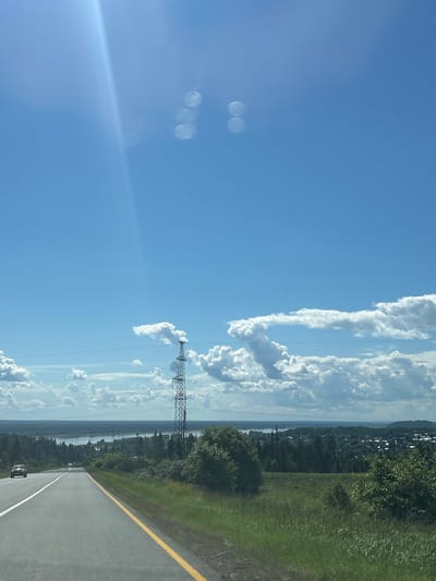 The image depicts a bright, sunny day with a clear blue sky and scattered white clouds. The sun is partially visible, casting a strong glare and creating lens flare artifacts in the sky. In the distance, a wide body of water, likely a river or lake, is visible, bordered by a lush green landscape. A power line tower stands tall on the right side of the frame, with power lines stretching across the horizon. The foreground features a paved road with a yellow line marking its edge, and on the left, a car is driving away from the camera. The environment suggests a rural or semi-rural setting, possibly near a town or village, consistent with the location of Нечкино, Russia, known for its scenic natural beauty and proximity to the Kama River. The overall impression is one of a serene and expansive natural vista on a summer day.