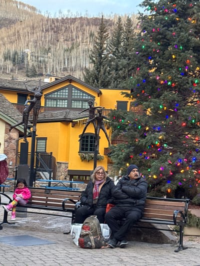Two adults sit on a park bench near a building painted yellow. A child wearing a pink jacket and striped tights is sitting on a bench nearby. A large tree with colorful lights decorates the scene. Bronze statues of figures in motion are placed in front of the building. A mountain range with leafless trees is visible in the background under a gray sky.