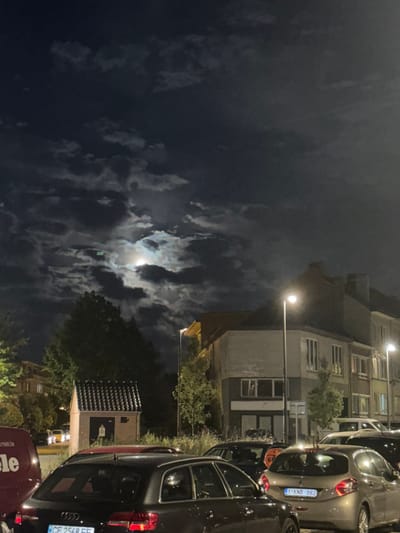 The image depicts a nighttime scene in Strombeek-Bever, Belgium, dominated by a cloudy sky with a bright moon partially visible through breaks in the clouds. In the foreground, several cars are parked, including a dark-colored sedan in the lower left and a lighter-colored hatchback with a license plate reading "1 KNB 982" to its right. A red car is partially visible behind the dark sedan. To the left, a portion of a red van with the word "Miele" is visible. In the mid-ground, there is a small, brick structure with a dark roof, possibly an electrical substation or utility building, situated amidst greenery. Behind this structure, trees are present. Further back, residential buildings with illuminated windows line a street, with streetlights casting pools of light. The overall atmosphere is nocturnal and somewhat atmospheric due to the dramatic cloud and moon formation.