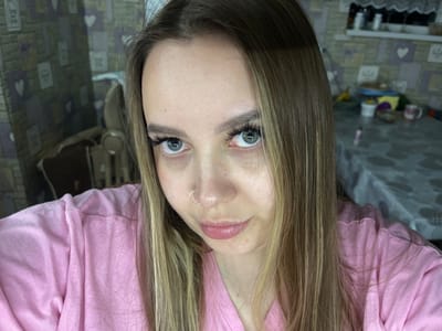 A portrait of a woman with blonde hair, wearing a pink shirt and a nose ring, oriented upside down. The background appears to be a room with a tiled floor and various objects scattered on a shelf. No location information pertaining to Port-Katon, Russia, is present.