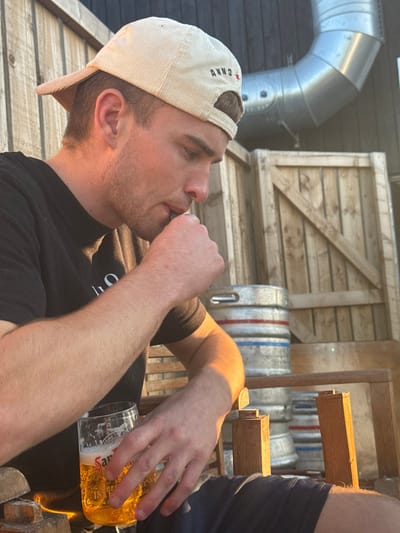 The image depicts a male individual, appearing to be in his twenties or thirties, seated in an outdoor setting at The Annos Arms, located at 15 Friarage St, Northallerton, DL6 1DP, United Kingdom. He is wearing a black t-shirt, dark shorts, and a light beige corduroy baseball cap with the brim backward, displaying "ANNO" and a red star embroidered on the rear.
The subject is illuminated by direct sunlight on his right side. His left hand holds a transparent glass containing a golden, bubbly liquid, identified by "San Mig" branding, consistent with San Miguel beer. His right hand is raised towards his mouth, holding a small, dark cylindrical object, from which he appears to be inhaling. He is looking downwards and to the right of the frame.
The background consists of vertical wooden slatted panels or fencing. To the right, a large, curved, metallic ventilation duct is visible, along with a wooden gate or panel. Multiple silver-colored beer kegs, some marked with red and blue bands, are stacked in the background, further indicating a pub or brewery environment. The seating is part of a wooden outdoor chair. The overall scene suggests a casual, outdoor pub garden setting under strong, natural, warm light.