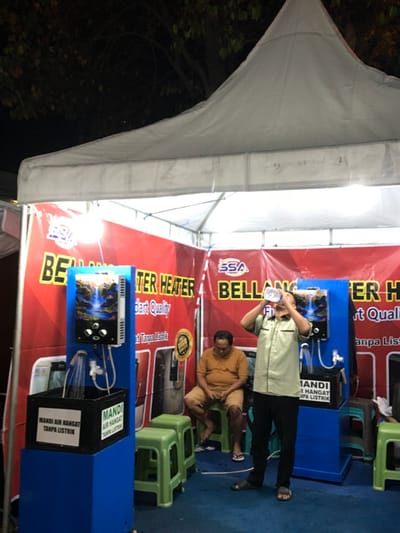 The image shows a booth in Kecamatan Batu, Indonesia, at coordinates (-7.866, 112.514). The booth features two water heaters displayed on blue stands with black trays and black boxes at the base. The booth is located under a white tent with a red and white banner that reads "BELANJA TER HEATER" with a smaller banner below that says "part quality" in English. There are three green plastic stools. A man in a light green shirt is standing in front of a water heater drinking from a bottle, facing towards the camera. A man in a brown shirt is seated on a green stool, holding a black object in his hands, and looking to the right. The booth is located in an outdoor setting under the tent with a dark background. The scene is well lit, with fluorescent lights illuminating the interior of the booth. There is a small amount of water splashing from a showerhead on one of the water heaters. The setting appears to be a promotional event for the water heaters.