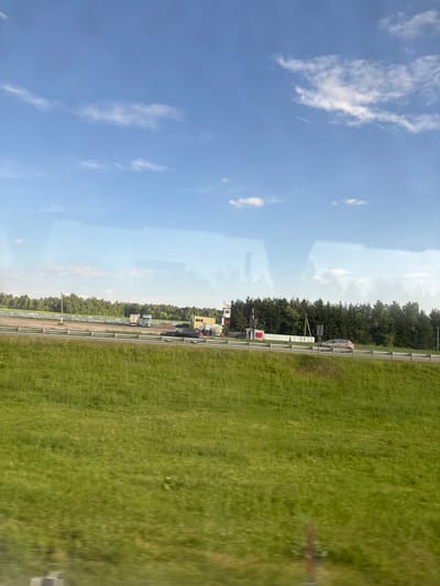 This is a daytime, eye-level, outdoor, long shot photograph taken from a vehicle, likely in motion, capturing a view of a highway and its surroundings under a clear blue sky with scattered white clouds. The foreground is dominated by a lush, green grassy field with some out-of-focus elements suggesting movement. Beyond the field, a highway runs horizontally across the frame, separated from the field by a metal guardrail.
On the other side of the highway, a row of trees forms a dense green backdrop. In front of the trees, several structures are visible. On the left side, a white truck is parked near a building. Further to the right, there's a yellow building with a white roof, a tall red and white tower, and a white fence. A dark-colored SUV is driving on the highway in the middle section of the image, and a lighter-colored sedan is driving on the highway further to the right. Two traffic lights are visible, one red and one green, positioned above the highway. There are also poles with traffic signs. The overall scene suggests a rural or semi-rural area with highway infrastructure. The image does not provide specific visual cues to pinpoint the location within Kazan, Russia.