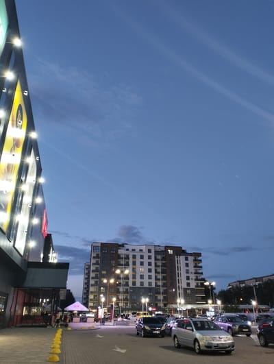The image shows a street scene in Ivano-Frankivsk, Ukraine, at dusk. The sky is a gradient of blue, with some clouds. On the left is a multi-story building with a brightly lit facade. A KFC restaurant is visible at the base of this building. In the foreground, yellow spherical bollards line the street, and an arrow is painted on the pavement indicating the direction of traffic. Several cars are parked or moving along the street. In the center of the frame, a tall residential building is visible, illuminated by streetlights. People are seen walking around the KFC entrance.