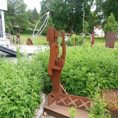 This photograph captures an outdoor sculpture park with several abstract metal sculptures set against a backdrop of lush green trees and manicured lawns. In the foreground, a prominent rust-colored sculpture, constructed from angular metal plates, stands amidst dense green foliage. This sculpture is positioned on a tiered metal base with an ornamental crosshatch design. To the left, a modern building with a white facade and large windows is partially visible, with a staircase and ramp leading up to its entrance. Further into the park, other sculptures can be seen, including a large, white, elliptical structure and several tall, thin metal forms. The scene appears to be set in a public space, possibly a park or a cultural institution, located in Eisenach, Germany. The weather is overcast, contributing to the subdued lighting. No people are clearly visible in the foreground, although a distant figure in a red jacket can be observed near a sign in the mid-ground. The overall impression is one of a contemporary art installation within a natural, landscaped environment.