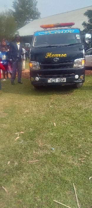 The image shows a black hearse van positioned on a grassy field in Esise ward, Kenya. The hearse displays "Hearse" in gold cursive text on its front grille and in blue cursive text on a light blue banner above the windshield. A red and amber emergency light bar is affixed to the vehicle's roof. The Kenyan license plate "KCV 304W" is visible on the front bumper. To the left of the hearse, a man in a dark suit jacket and a yellow bow tie stands, facing slightly left with a subtle smile. Other individuals, including what appear to be children, are partially visible behind him. The immediate ground beneath the vehicle is dirt, transitioning to sparse green grass in the foreground. In the background, a building with a light-colored roof is present, surrounded by trees under an overcast sky. The scene appears to be an outdoor gathering.