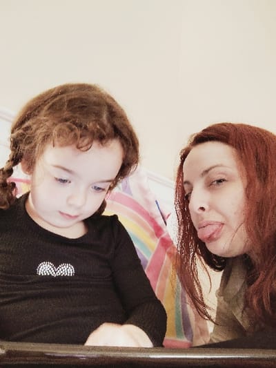 The image shows two women in an indoor setting. The woman on the left is a young girl with long brown hair in a braid, wearing a black long-sleeve shirt. The woman on the right has long red hair and is wearing a light-colored shirt. The girl is looking down at a tablet or laptop. The woman is looking at the camera and sticking her tongue out. There is a colorful rainbow-striped blanket in the background. The location of the image is in South San Francisco, United States, based on the coordinates 37.66 latitude and -122.412 longitude. The image captures a casual and informal moment between two people, possibly mother and daughter, in a domestic setting.