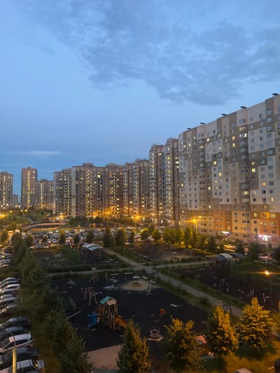 The image depicts a view of a residential area in Domodedovo, Russia, during twilight. Dominating the scene are multiple tall, multi-story apartment buildings, their windows illuminated, indicating occupancy. In the foreground, a children's playground is visible, featuring various play structures such as swings, slides, and climbing equipment, with a sandpit in the background. Adjacent to the playground is a parking lot filled with numerous cars, suggesting a densely populated urban environment. Trees and green spaces are interspersed throughout the area, providing a contrast to the concrete structures. The sky is overcast with a gradient of blues and grays, typical of the transition from day to night. The lighting suggests that artificial streetlights are also present, casting a warm glow on some of the pathways and play areas. There are no discernible individuals in the image engaged in any specific actions.