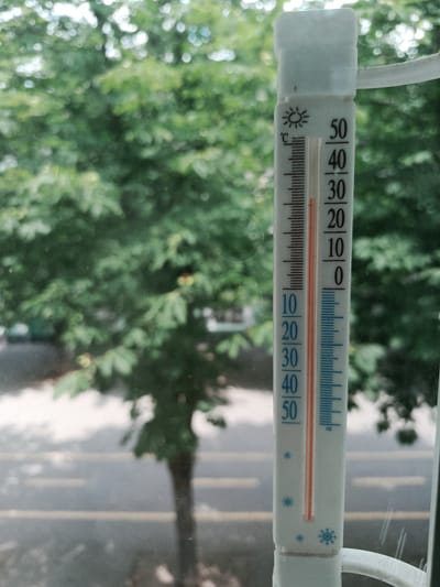 An outdoor thermometer is shown mounted on a window, with a blurred background of green foliage and a road with yellow lines. The thermometer displays a temperature between 40 and 50 degrees Celsius, with the red liquid column reaching approximately the 46-degree mark. The thermometer has markings for Celsius, with a sun icon at the top and snowflake icons towards the bottom. The scene appears to be a residential area in Pokrov, Ukraine, possibly a balcony or window of an apartment building overlooking a street. There are no people or animals visible in the image. The lighting suggests it is daytime.