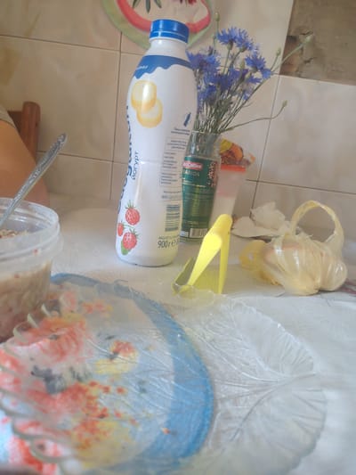 The image depicts a close-up, low-angle view of a tabletop setting, likely within a domestic environment in Коханаўскі сельскі Савет, Belarus. The primary subject is a partially visible bottle of yogurt with a blue cap and a white label featuring illustrations of strawberries and banana slices. Next to the yogurt bottle is a small glass vase containing a bouquet of blue cornflowers, some of which have buds.
To the left of the yogurt bottle, a clear plastic container holds a beige-colored food item, possibly oatmeal or porridge, with a silver spoon resting inside. In the foreground, a transparent, textured plate with a blue rim is visible, showing remnants of colorful food or decorations.
To the right of the yogurt bottle and cornflowers, a yellow plastic utensil, possibly a food clip or dispenser, is positioned. Further to the right, a translucent yellow plastic bag, partially filled and tied at the top, contains a light-colored, possibly food-related, item. A few crumpled white napkins are also present in the background. The overall scene suggests a casual meal or snack preparation, with a rustic and simple aesthetic. The background consists of tiled walls, suggesting a kitchen or dining area.