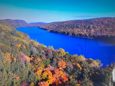 The image shows an aerial view of a long, narrow body of water, possibly a lake or reservoir, surrounded by forested hills. The water is a deep blue color. The hills are covered in trees displaying a mix of autumnal colors, including green, yellow, orange, and red. There are mountains in the background. The sky is a clear light blue. There are no people visible in the image. The location is unspecified, but given the fall foliage and topography, it is likely a temperate region. It is not known to be Basingstoke and Deane, United Kingdom.