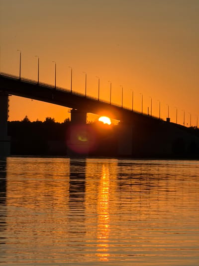 The image depicts a sunset over a bridge, likely the Kambarka Bridge, in Kambarka, Russia. The sun, partially obscured by the bridge structure, is low on the horizon, casting a warm orange glow across the sky and the water. The bridge, silhouetted against the vibrant sunset, is lined with streetlights. The water in the foreground reflects the colors of the sky and the bridge. Trees are visible in silhouette along the far bank of the river.