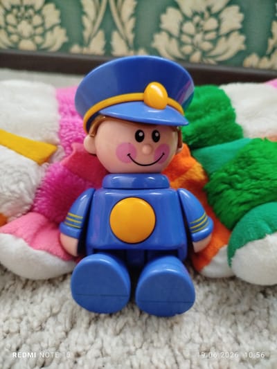 A plastic toy figurine of a smiling uniformed person, possibly a police officer or pilot, is positioned centrally in the frame. The figurine is blue and yellow with black eyes and pink cheeks. It is seated on a textured beige carpet, with colorful plush toys forming a backdrop. The background also includes a patterned wallpaper and a dark horizontal surface above it. The scene appears to be indoors, likely a domestic setting. The image is dated "19.06.2026 10:56" and tagged "REDMI NOTE 13" in the bottom left and right corners respectively. There is no visible indication of the location in the city of Асан-Чек, Kyrgyzstan.