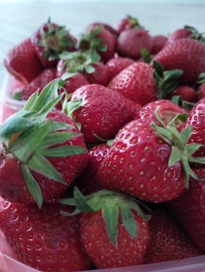 A close-up, high-angle shot shows a container overflowing with fresh strawberries. The strawberries are vibrant red with green stems and yellow seeds. The container appears to be transparent plastic, possibly a store-bought or reusable container. The background is slightly blurred, suggesting a table or countertop with a light-colored surface. The location in Krykhivtsi, Ukraine is not evident from the image alone.