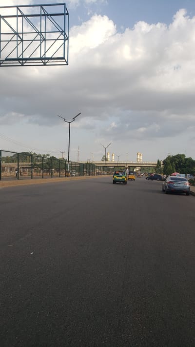 The image depicts a street scene in Hayin Banki, Nigeria. A four-lane road is visible, with a green and yellow auto rickshaw ("keke napep") traveling in the right lane. Cars are parked further down the road. The "Kaduna" structure with dome-like architectural elements is visible in the background. A billboard structure is visible on the left side of the road. Streetlights are along the side of the road, and there is fencing between the road and the sidewalk, separating it from the building structures. The sky is cloudy.