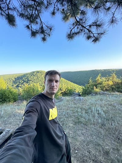 A young man with short brown hair, looking directly at the camera, is taking a selfie outdoors. He is wearing a black long-sleeved shirt with a yellow graphic on the chest. He is positioned on a grassy, rocky embankment overlooking a vast, densely forested valley under a clear blue sky. Pine branches with needles frame the top left of the image, obscuring some of the sky. The sun appears to be low in the sky, casting a warm light on the scene, suggesting it is either early morning or late afternoon. The overall atmosphere is peaceful and serene, with a sense of natural beauty and solitude. The location is identified as Pryvorottia Druhe, Ukraine.