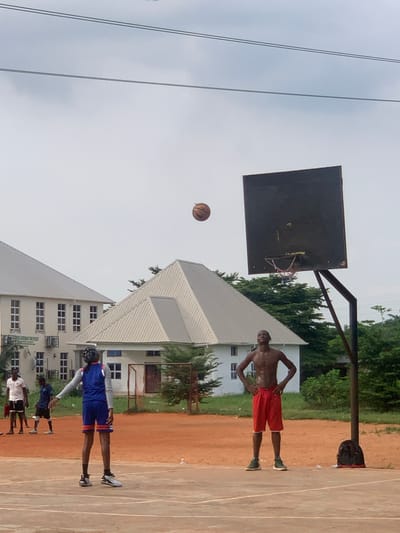 In Ukukwa, Nigeria, on an outdoor basketball court with a reddish-brown dirt surface, a group of young men are engaged in a game. The sky is overcast. The court is bordered by some trees and two large, white buildings with grey, gabled roofs. One of the buildings has multiple windows arranged in rows. The other building has a single, pitched roof. A basketball hoop with a black backboard and a red, white, and blue net stands on the right side of the frame. A black backpack rests on the ground near the base of the hoop.
In the foreground, two individuals are the primary subjects. On the left, a person wearing a blue and orange basketball uniform, complete with a bandana on their head, faces away from the camera, their left arm extended. To the right, a shirtless young man with muscular build stands with his hands on his hips, wearing red basketball shorts and green sneakers. A basketball is in mid-air above and to the left of the hoop, suggesting it has just been shot. In the background, on the left side of the court, two other young men are visible. One is wearing a white t-shirt and black shorts, and the other is in a blue t-shirt and dark shorts. They appear to be observing the game.