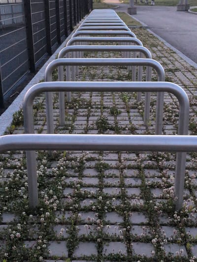 The image shows a row of metallic bicycle racks set against a backdrop of a dark metal fence. The ground beneath and around the racks is paved with rectangular concrete blocks, with a substantial amount of clover and small white flowers growing between them. The scene appears to be an outdoor urban environment, likely a public space like a park or a sidewalk. The lighting suggests it might be late afternoon or early evening. The overall impression is one of a functional urban amenity integrated with natural elements.