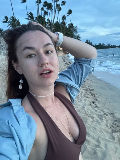 A woman is taking a selfie on a beach in Tong Yang, Thailand. She is wearing a brown bikini top and a light blue shirt, with her hand on her head. The background shows palm trees and the ocean. The sky is cloudy and the lighting suggests it is either dawn or dusk. There are small waves breaking on the shore, and the sand is light brown.