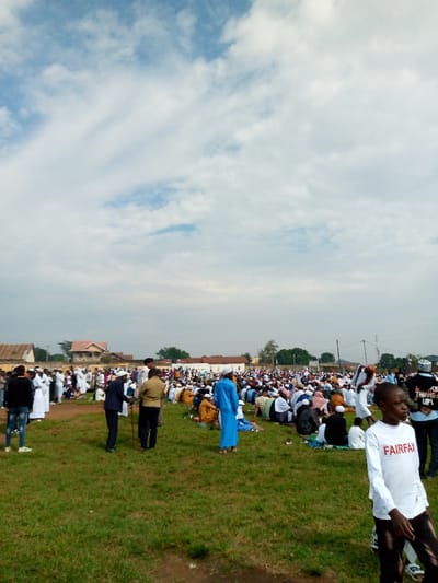 The image shows a large gathering of people in an open grassy area, likely a field or park. The crowd is predominantly male and appears to be attending a religious ceremony or gathering. They are dressed in traditional clothing, with many wearing white robes and some wearing prayer caps. The event is taking place in Beni, Democratic Republic of Congo, at the coordinates provided. The weather is sunny with a clear blue sky and scattered white clouds. The scene is observed from a distance, the focus is on the crowd, and the image is taken from a slightly elevated position. The crowd is spread out across the grassy area, with some people standing and others seated in rows. There are a few trees and buildings in the background.