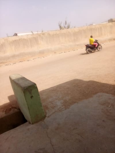 The image depicts an outdoor scene in Tudun Murtala, Nigeria, under clear, bright conditions. In the lower left foreground, a light green rectangular concrete block stands vertically, casting a dark shadow to its right. It partially borders a visible drainage opening or channel in the concrete ground. The immediate ground surface is a cracked, dirt-stained concrete slab.
Beyond the foreground, a wide, light-colored dirt road extends from left to right. A single individual is observed riding a dark-colored motorcycle from left to right across this road. The rider is seen from the rear, wearing a bright yellow shirt or top and reddish-pink trousers.
In the background, a long, light brown or beige-colored concrete wall, showing vertical discoloration marks, stretches horizontally across the frame. Above the wall, a few sparse, light-colored trees or bushes are visible, and parts of a white building can be seen behind the wall against a pale sky. Strong sunlight illuminates the scene, creating distinct shadows on the ground.