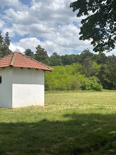 The image depicts a small, white, one-story building with a red tile roof situated on a grassy area. A forest is visible in the background. The sky is partly cloudy. The location appears to be an outdoor setting within the Sopot Urban Municipality, Serbia. There are no people visible in the image.