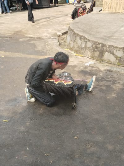 The scene depicts a man kneeling on a paved surface, possibly a street or courtyard, in Pendem, Indonesia. The man, wearing a black jacket, black pants, a red headband, and blue sneakers, is hunched over a large, black and red woven object with horsehair-like fringe. The object appears to be a decorative mask. A few individuals are visible in the background, along with a speaker and a small structure made of bamboo. The ground is slightly wet.