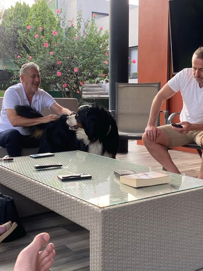 Two men are outside, one standing over a table and the other sitting. A large Bernese Mountain Dog stands between them. The standing man holds a phone. The sitting man is petting the dog. There is a glass-topped wicker table with several remote controls and a book on it. In the background are plants and part of a building. A pair of feet are visible in the bottom right.