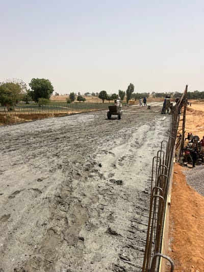The image depicts an active construction site in Gyellesu, Nigeria, under a clear, bright sky. The central subject is a wide, newly poured concrete surface, likely forming a road or bridge deck, extending from the foreground into the midground. The concrete appears wet and fresh, displaying numerous boot and tire track impressions. In the midground, a worker pushes a two-wheeled wheelbarrow containing material across the wet concrete. Further beyond this worker, several other individuals are visible on the concrete surface, actively engaged in construction work, possibly spreading or leveling the material. Along the immediate right edge of the concrete slab, exposed vertical and horizontal rebar structures are visible, indicating ongoing reinforcement and formwork. Below this right edge, on a lower earthen level, multiple workers are clustered around piles of grey aggregate (gravel), with some appearing to operate equipment or handle materials. The background features a rural landscape with sparse green trees and shrubs interspersed with dry, open fields.