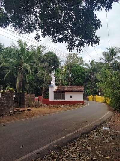 The image shows a road in Marna, India. The road curves to the left and is lined with vegetation. A small white building with a red border and tile roof is on the right side of the road. A yellow building is further up the road. Power lines are visible overhead. A stone fence and gate are in the foreground. Trees, including palm trees, surround the area.
