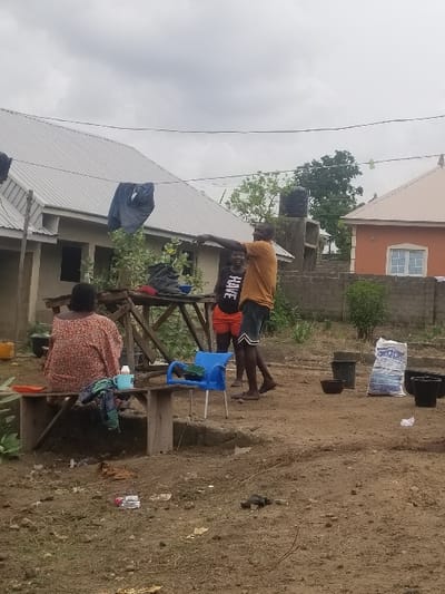 The image depicts a scene in the backyard of a home located at coordinates 7.81 latitude and 6.704 longitude in Pati Ridge, Nigeria.  A woman wearing a patterned, pink and brown dress sits on a wooden bench, holding a blue cup in her right hand. There is a blue plastic chair next to her, and a pair of blue sandals lying on the ground beside it.  A small, white plastic bag is visible at the base of the wooden bench.  A second woman, wearing a pink shirt that reads "FIVE", red shorts, and dark sandals, stands behind the bench. She is talking to a man, who is wearing a brown shirt, blue jeans, and black sandals.  The man is pointing to something out of frame with his left hand.  The man and woman are standing in front of a small wooden table, where a bag of fertilizer is sitting. A black pot and a black bucket are visible in the background, as is a grey, concrete wall.  The ground is dusty and uneven, and there is foliage growing in the background.  The scene is captured outdoors, on a cloudy day, with an overcast sky.